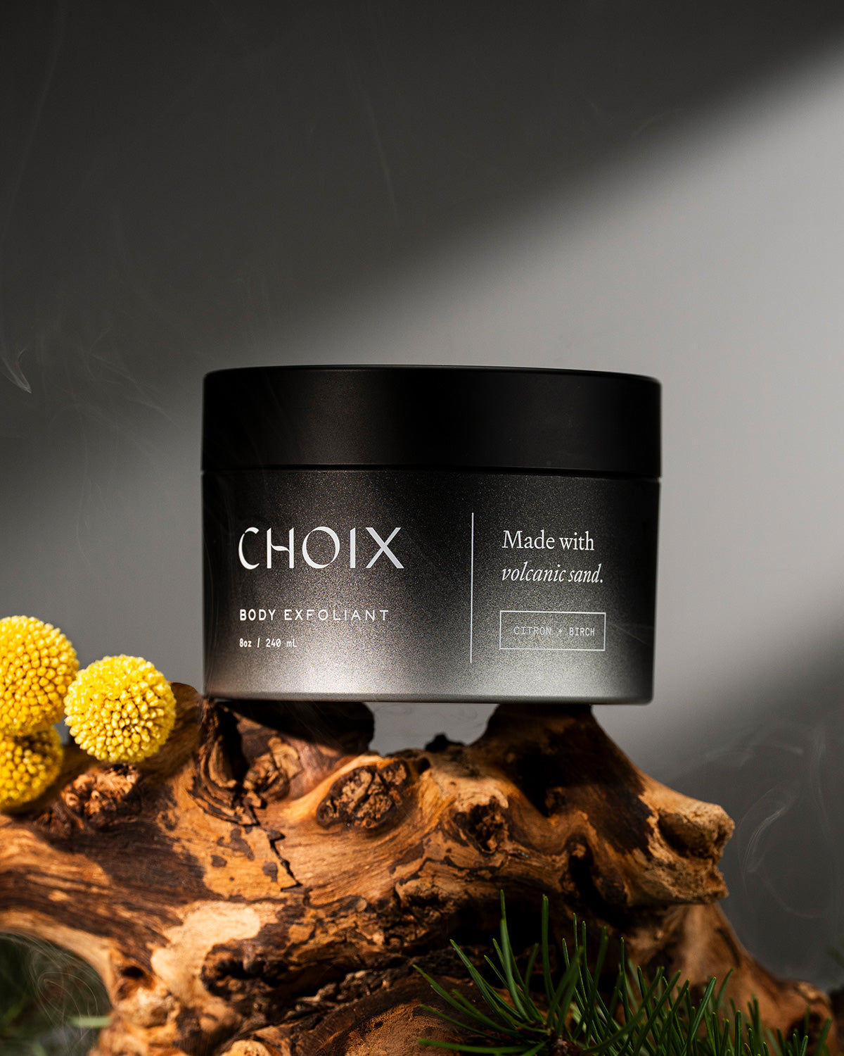 Black jar of CHOIX body exfoliant on a birch tree brand with yellow flowers.