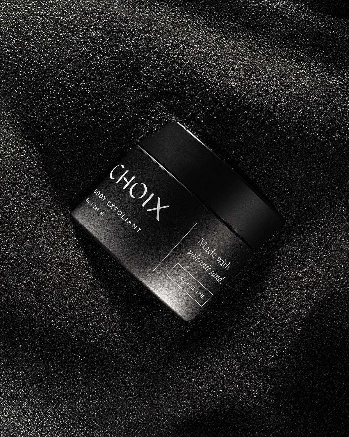 Black jar of fragrance free 'CHOIX' body exfoliant on a volcanic sand background